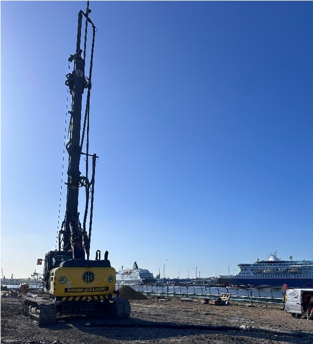 Photograph of a construction site near a waterfront with a large yellow piling rig in the foreground. A cruise ship and smaller boats are visible in the background under a clear blue sky.