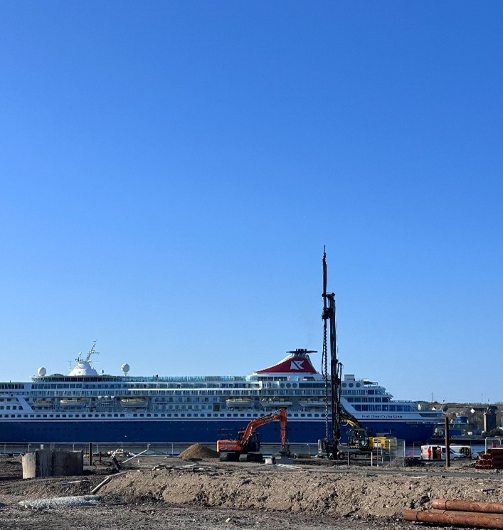 Photograph showing a large cruise ship docked near a construction site with a piling rig and piles of dirt in the foreground.