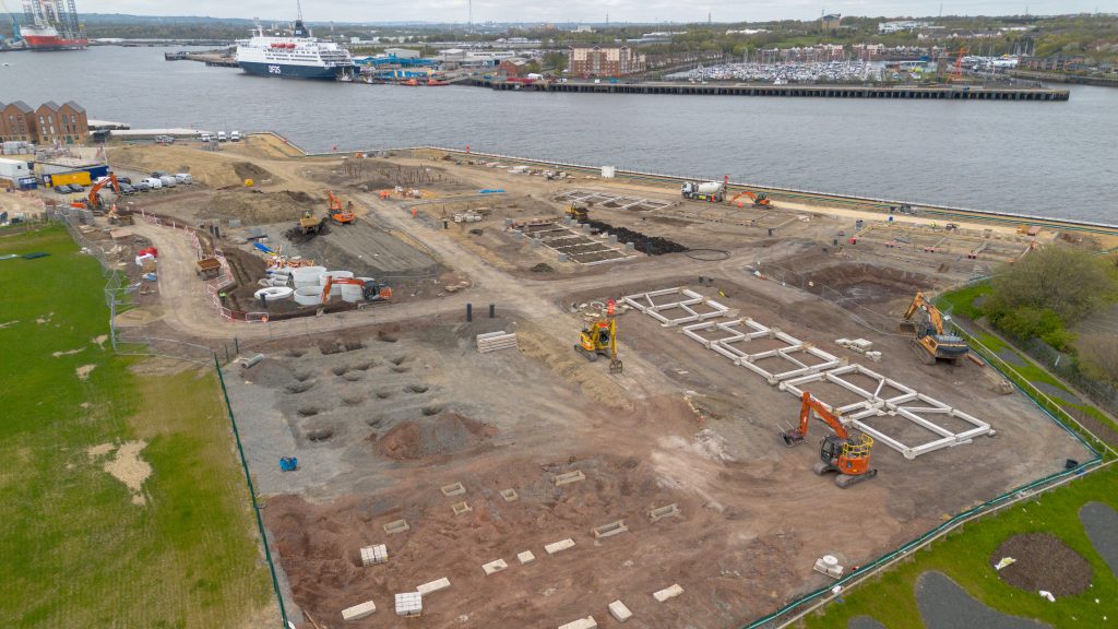 Aerial photograph of a large construction site near a waterfront, showing precast foundation work in progress.